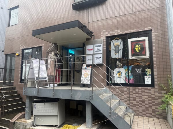 一棟建築的入口，金屬樓梯旁掛著數塊店家招牌，櫥窗展示著印花 T 恤